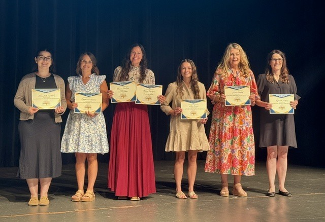 Six female Harrison School District teachers stand side-by-side on the auditorium stage, proudly holding up the official Harrison Public Schools Foundation grant certificates they received for their classroom projects.