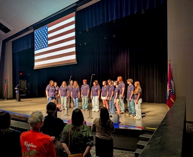Harrison choir students are singing the National Anthem for the school district back to school pd. The American flag hangs in the backdrop.