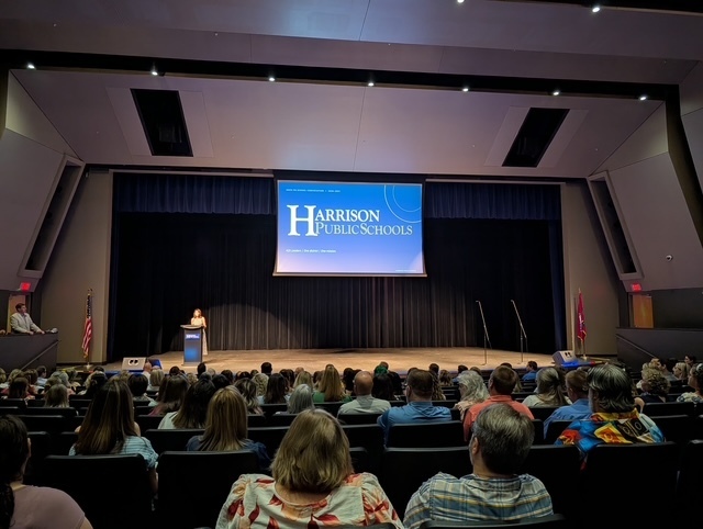 View from the back of the auditorium showing rows of Harrison School District staff seated and looking toward the stage. A speaker stands at the podium beneath a large screen reading "Harrison Public Schools."