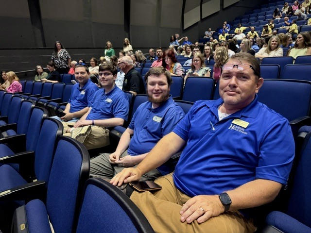 Four male Harrison School District staff members sit in a row of blue auditorium seats, smiling for the camera. They are wearing matching bright blue polo shirts embroidered with the "Harrison Public Schools" logo and nametags, sitting alongside other staff members in the audience before the presentation begins.
