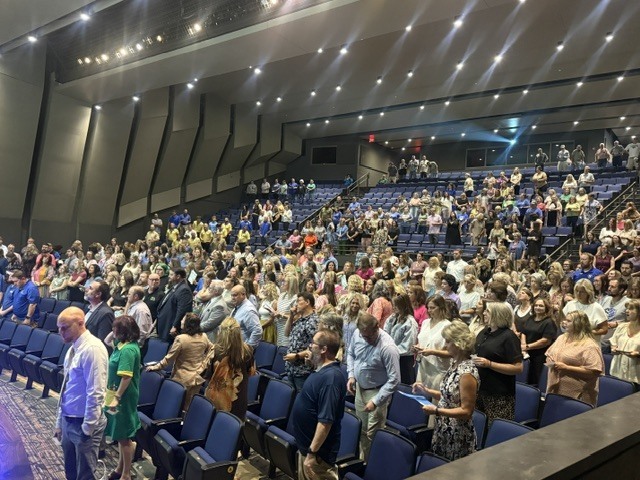 A wide, elevated side angle of the Performing Arts Center auditorium filled with Harrison staff members standing at their seats during the morning program.