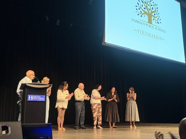Wide shot of the stage at the Harrison Performing Arts Center. Members of the Harrison Public Schools Foundation board stand in a line on stage, smiling and applauding next to a podium. A projector screen displays the "Harrison Public Schools Foundation" logo.