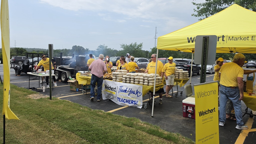 An outdoor setup in a parking lot where representatives from Weichert, REALTORS® - Market Edge, dressed in matching yellow shirts, serve lunch to Harrison Schools staff. The area features a large mobile smoker grill, a yellow pop-up tent, and tables covered in yellow tablecloths filled with stacked boxed meals. A banner displayed across the main table reads "Weichert REALTORS Market Edge - Thank You TEACHERS!"