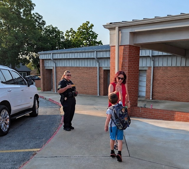 A young female student with a backpack high-fives a male school administrator in a white shirt and tie outside a school brick entryway on a sunny morning.