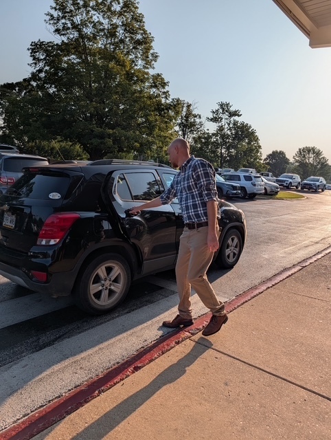 A male staff member in a plaid shirt and khaki pants opens the back passenger door of a black SUV to assist a student in the school drop-off line.
