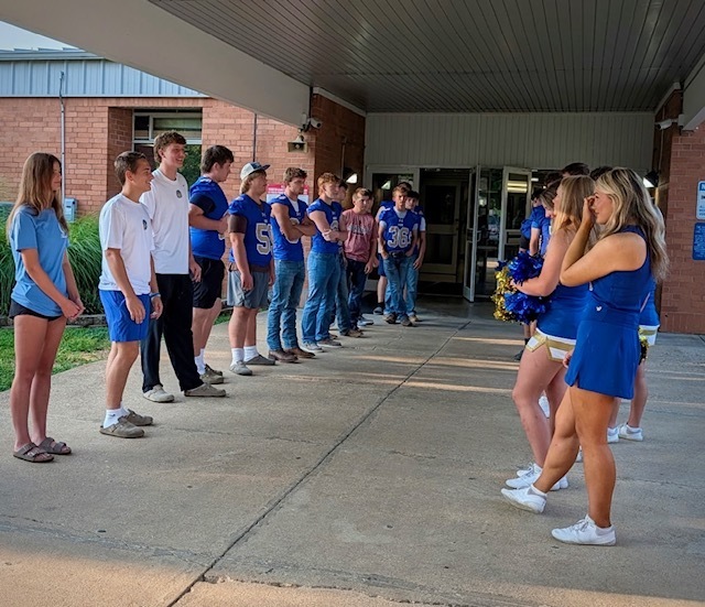 A student carrying a backpack walks along the sidewalk next to a line of yellow school buses parked in front of a grassy hill.