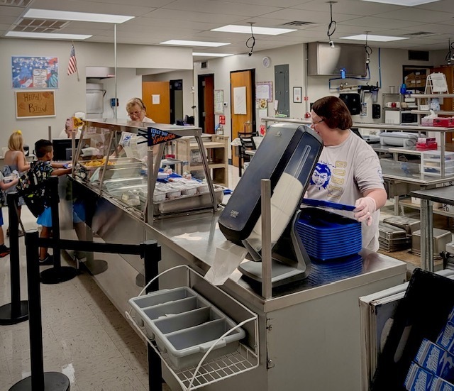 Smiling cafeteria staff members stand behind the serving counter in a clean, commercial school kitchen, preparing to serve meals to students.