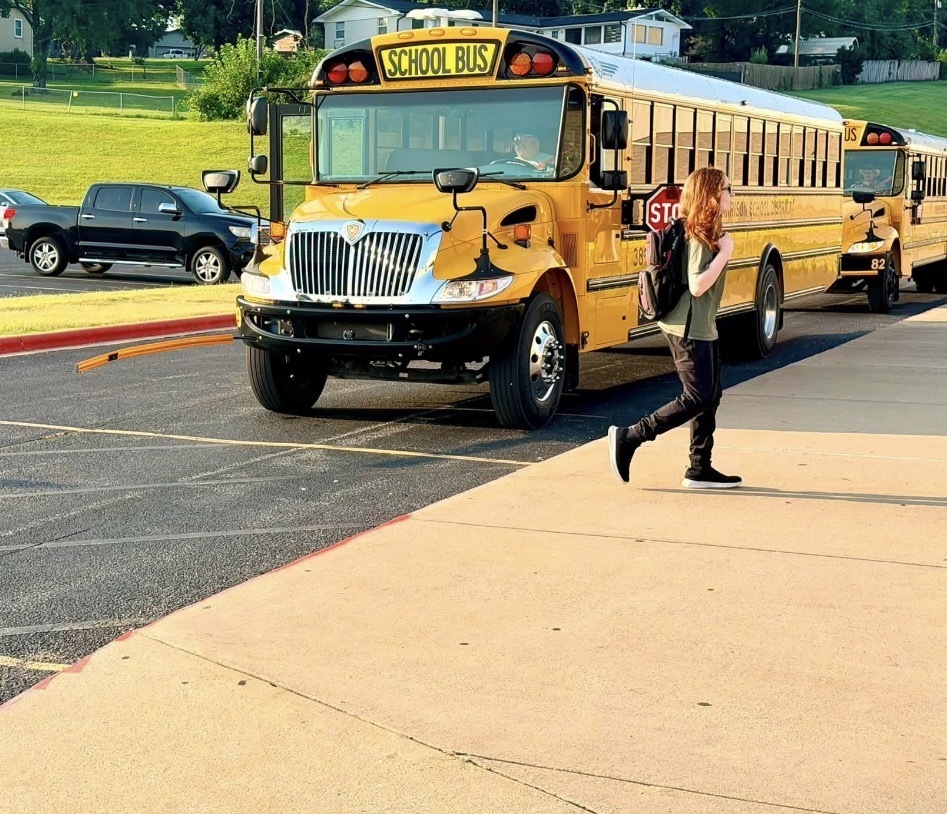 A young boy wearing a backpack high-fives a female staff member in a red top at a school drop-off line, while a female police officer smiles in the background.