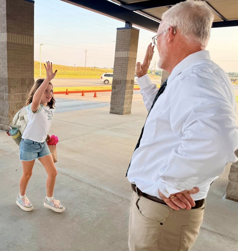 A young female student with a backpack high-fives a male school administrator in a white shirt and tie outside a school brick entryway on a sunny morning.