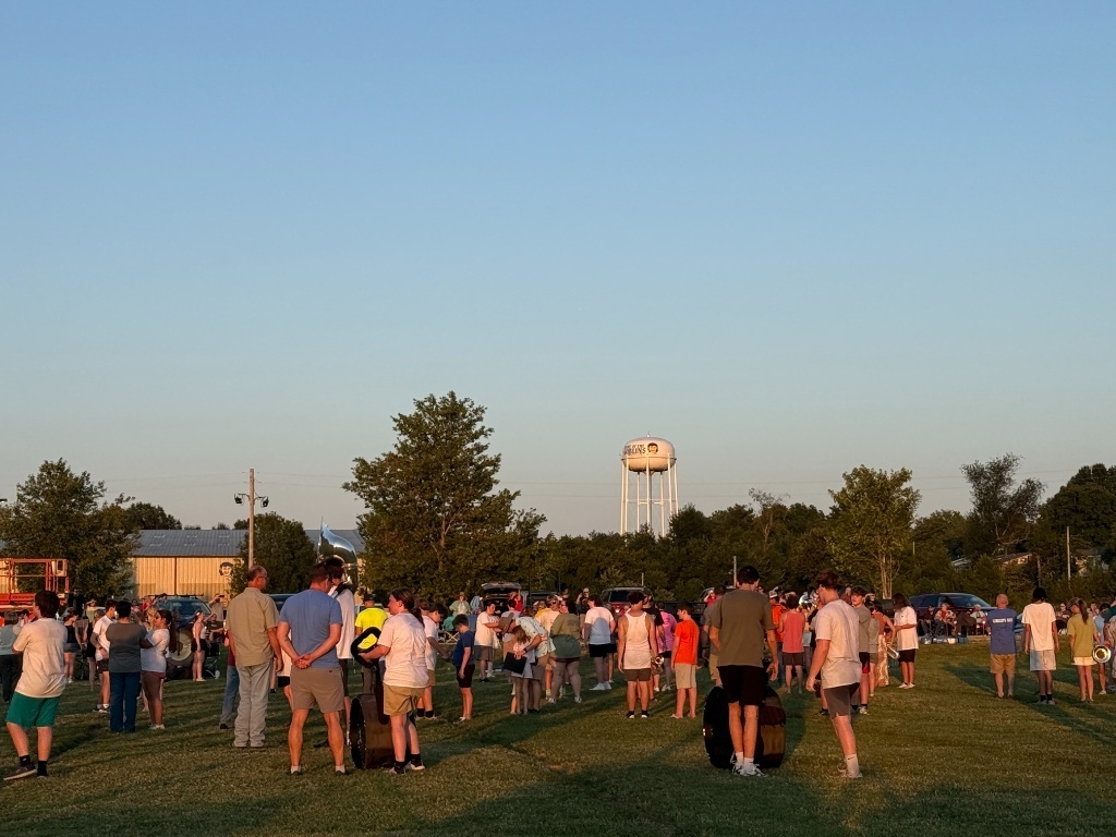 A wide outdoor shot of the Harrison Band students and a crowd of family members gathered on a grassy practice field under a clear blue sky. In the background, a white water tower with "Home of the Goblins" and a mascot logo stands tall beyond the trees.