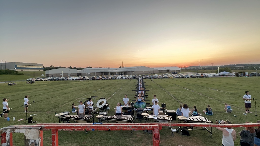 high-angle view looking down at the marching band front ensemble (pit) percussionists set up with marimbas and xylophones on the grass field. A long line of color guard members stretches straight back into the distance under a bright sunset sky.