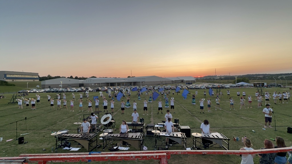 A high-angle, wide shot of the full marching band spread out in formation on the practice field during the sunset performance. Front ensemble instruments are lined up in the foreground, color guard members hold blue flags in the center, and brass and woodwind players are lined up across the field.