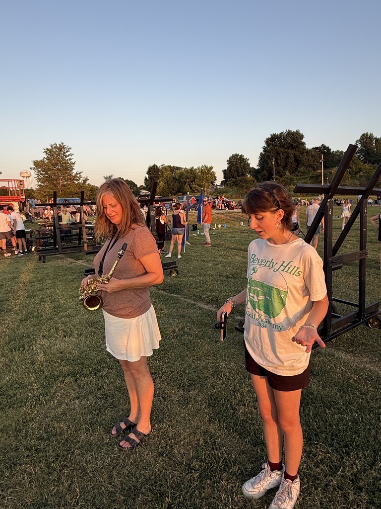 A woman holding a saxophone and a young girl standing next to her on the grass field, both bathed in the warm, golden light of the setting sun. Other event attendees and equipment stands are visible in the background.