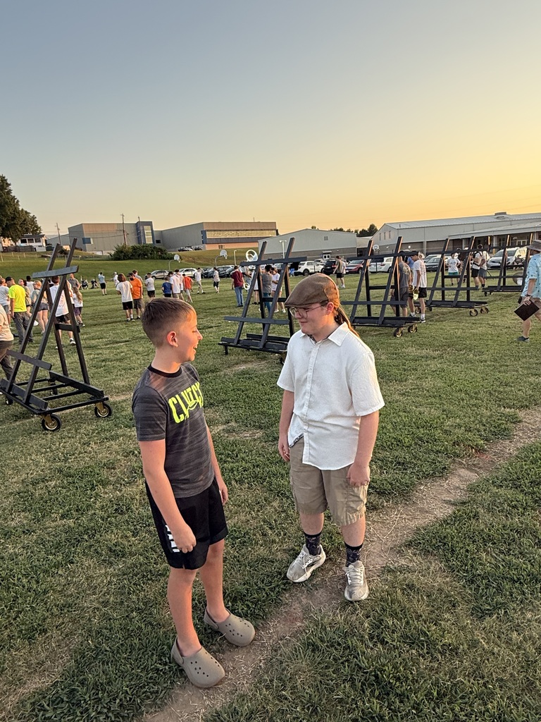 Two young boys standing on the grass field and smiling while talking to each other. One boy wears a dark athletic t-shirt and shorts, while the other wears a white short-sleeved button-down shirt, shorts, and a brown flat cap. Black equipment carts are parked in the background.