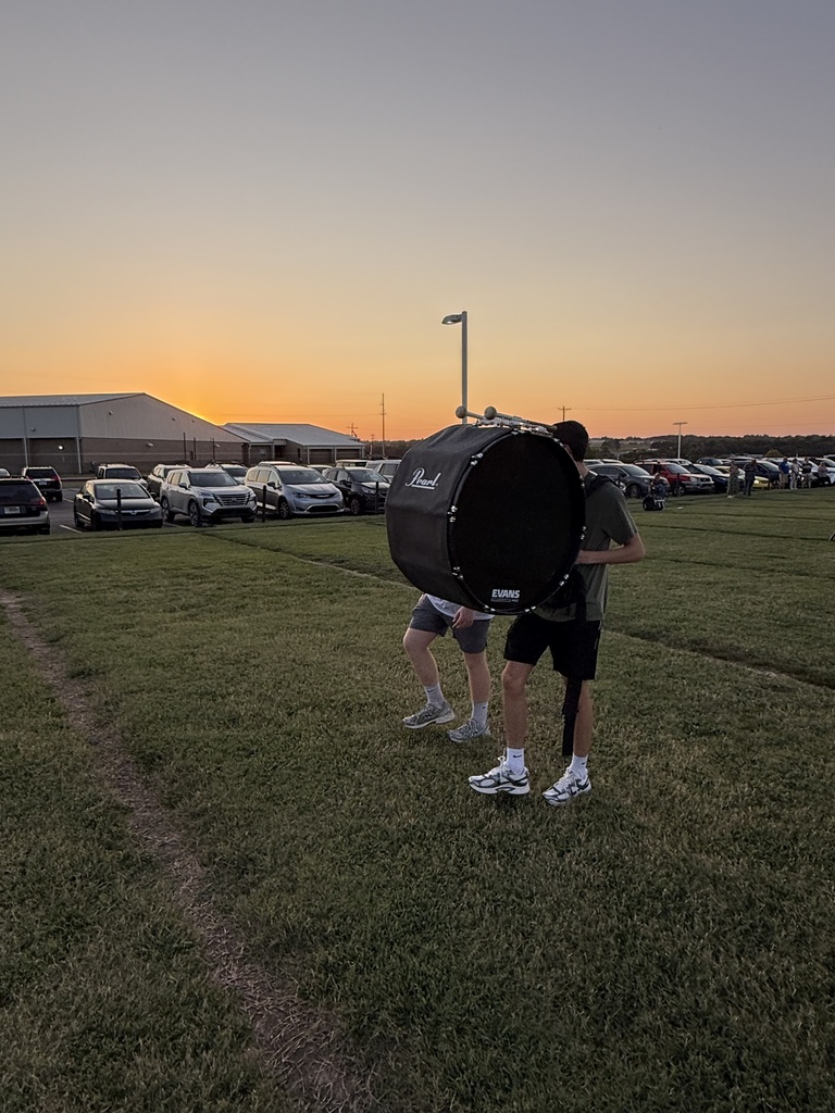 Two band students walking across a grass field at sunset. The student in the foreground is carrying a large, black Pearl marching bass drum on his back, silhouetted against a vibrant orange and yellow sunset sky.