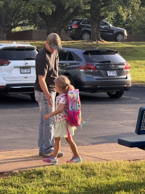 A man in a baseball cap and t-shirt greets a young school girl with a colorful Minnie Mouse backpack as she walks along the school sidewalk.