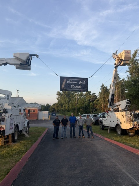ive Entergy workers stand in a row on a school driveway under a large black banner suspended between two utility bucket trucks that reads, "Welcome Back Students."