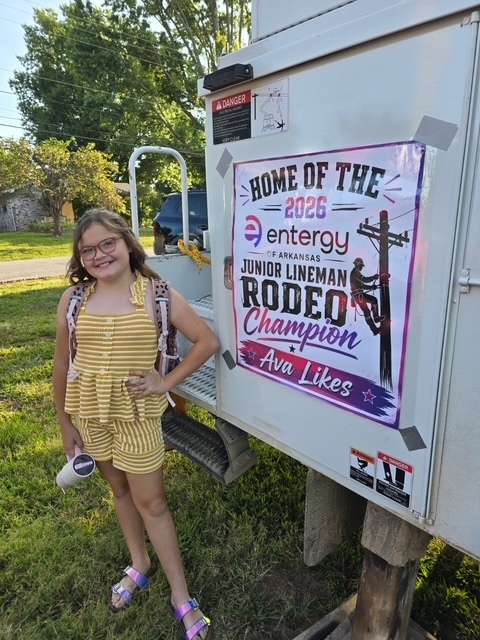 A smiling young girl with glasses and a striped yellow outfit stands proudly next to a white Entergy truck featuring a large poster that reads: "Home of the 2026 Entergy of Arkansas Junior Lineman Rodeo Champion Ava Likes."