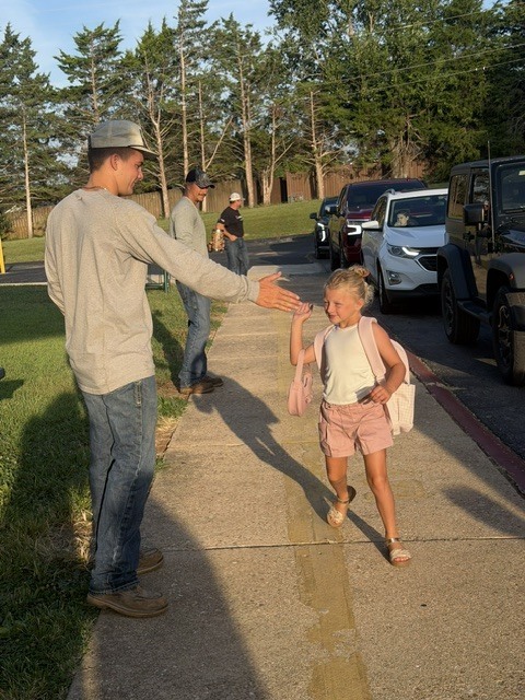 .A young man wearing an Entergy long-sleeve shirt, jeans, and a baseball cap leans down on a school sidewalk to offer a friendly high-five to a smiling young girl wearing a pink outfit and a backpack.