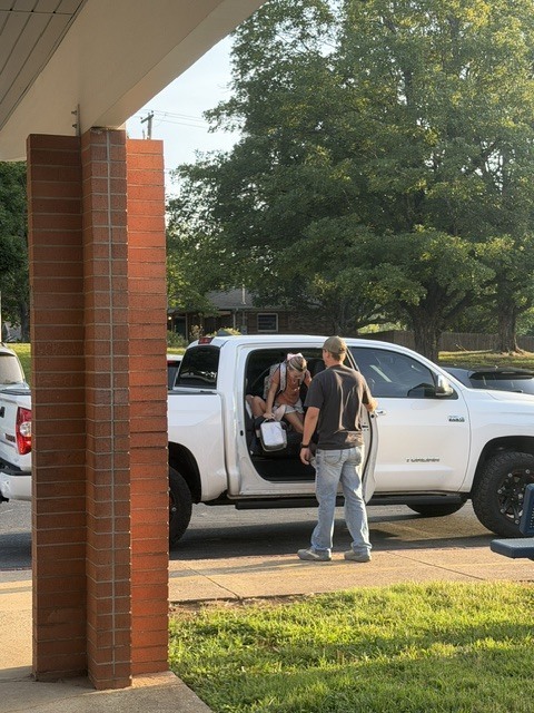 man stands by the open door of a white pickup truck, helping a young school girl gather her backpack and things during morning drop-off.