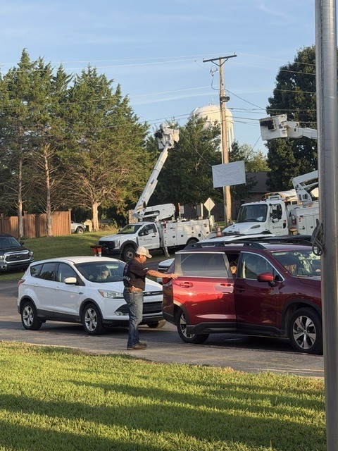 A line of cars during a school morning drop-off, with two Entergy utility bucket trucks parked in the background and a worker assisting near a vehicle door.