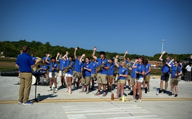 A wide shot of the Harrison High School pep band standing on a newly paved concrete bridge under a clear blue sky. The students, wearing matching blue t-shirts and khaki shorts, are holding instruments and waving enthusiastically toward the camera. A band director in a blue shirt and khaki pants stands facing them with his arm raised.