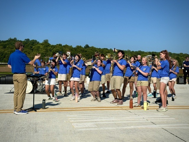 Members of the Harrison High School band playing their instruments on a concrete bridge during a sunny day. Students in matching blue t-shirts and khakis are actively playing trumpets, saxophones, and flutes, while the band director conducts from the left side of the frame.
