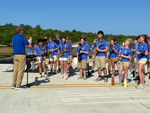 A close-up shot of the Harrison High School pep band performing on a wide concrete road. The student musicians are wearing matching blue shirts and khaki shorts while playing saxophones, clarinets, and brass instruments. The band director stands on the left, conducting the ensemble under a bright, sunny sky.