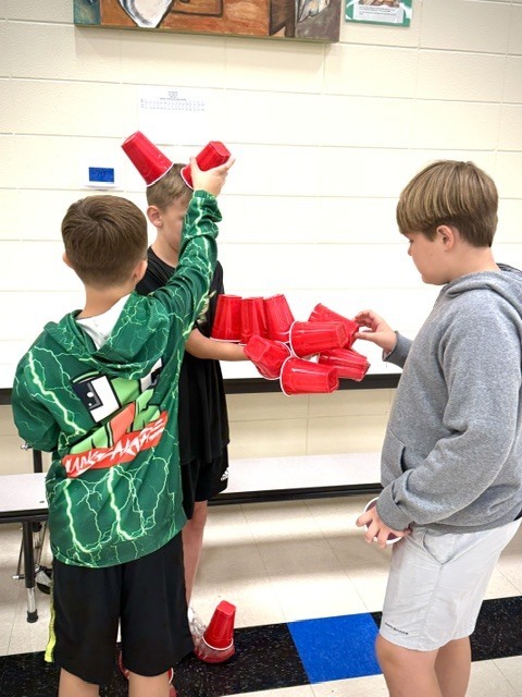 Three boys stand around a school table participating in a fun cup-stacking challenge. One boy wears a bright green lightning-bolt hoodie, while the others carefully balance a growing structure of red plastic cups.
