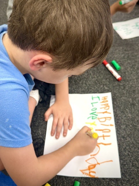 A close-up view from above of a young boy with short brown hair laying on the carpet. He is using a yellow marker to carefully write "Happy Birthday I LOVE you" in bright colors on a large white piece of paper.