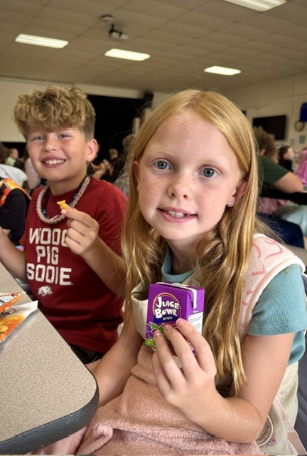 A smiling young girl with long strawberry-blonde hair and freckles holds up a purple grape juice box during snack time. In the background, a boy wearing a red shirt smiles for the camera while holding a small snack cracker.
