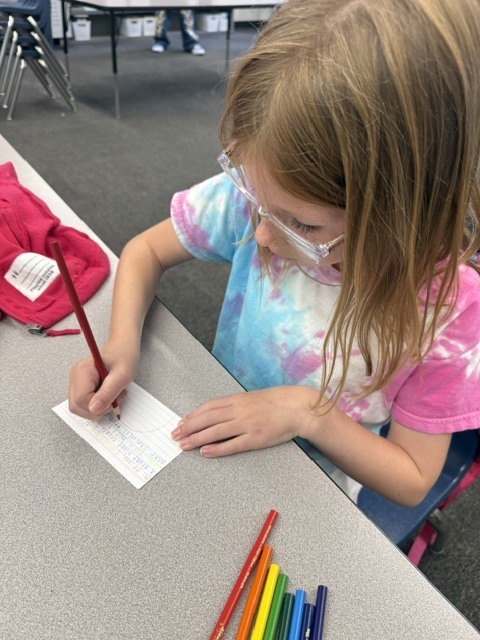 A young girl with long blonde hair and glasses sits at a gray desk, wearing a blue and pink tie-dye shirt. She is focused on writing a kind message with a colored pencil on a small index card. Several colored pencils are lined up neatly on the table in front of her.