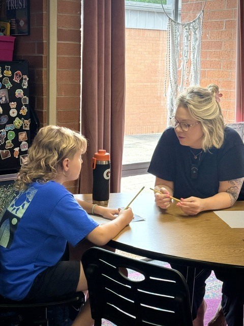 A female teacher with blonde hair and glasses sits at a wooden table, gently guiding and talking to a student with wavy blonde hair. They both hold pencils as they work through a lesson together in a bright classroom.