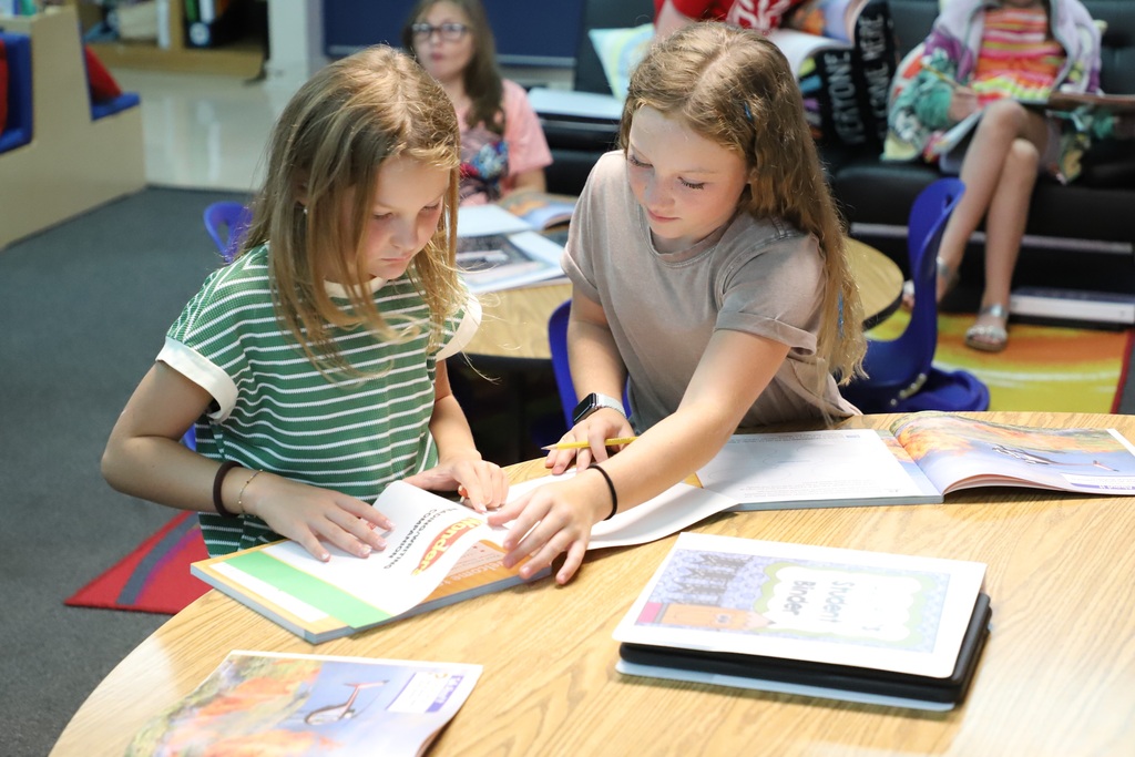 Two young girls leaning over a wooden table, happily collaborating and turning the pages of an open physical binder and workbook together during a lesson.