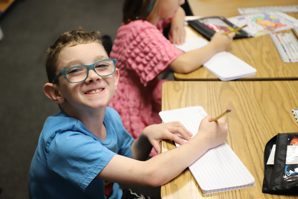 A young boy with glasses and a blue shirt smiles brightly at the camera while sitting at a wooden desk, holding a pencil and writing in a lined notebook.