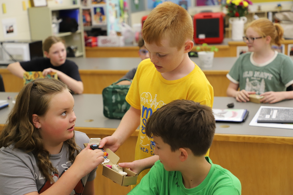 A group of four students collaborating at a classroom table. A boy in a yellow shirt shows a small homemade cardboard project to a girl in a grey shirt, while another girl and boy interact in the background.