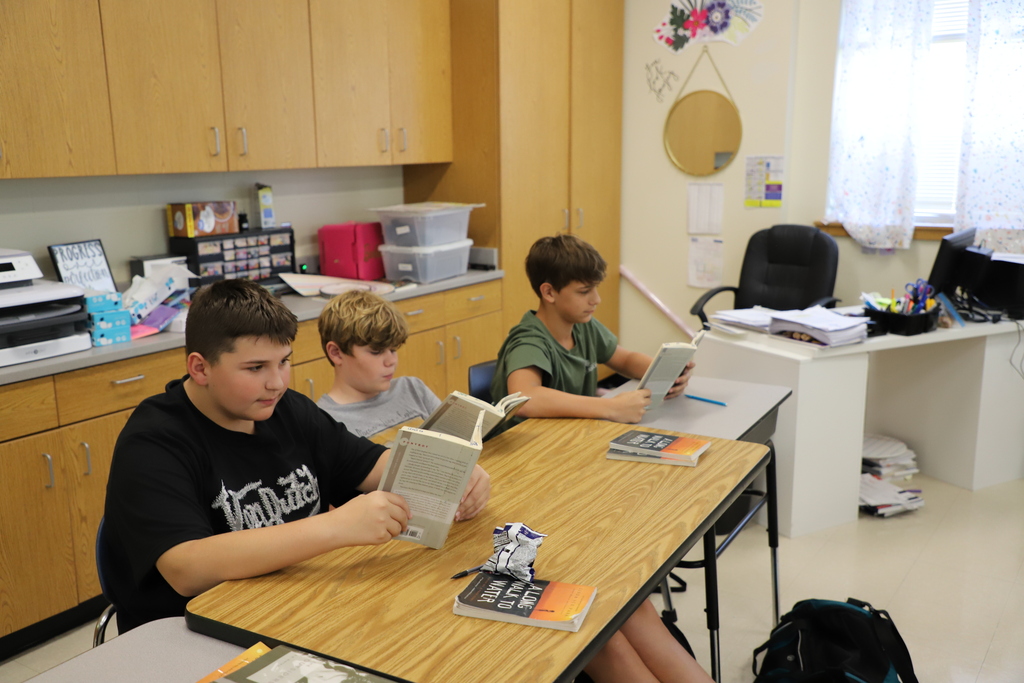 Three school boys sitting at long wooden classroom desks, deeply focused on reading physical paperback books during a quiet study session.