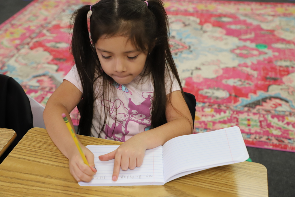 A young girl with her hair in twin pigtails sits at a school desk, using her finger to track her place as she writes with a pencil in a notebook.