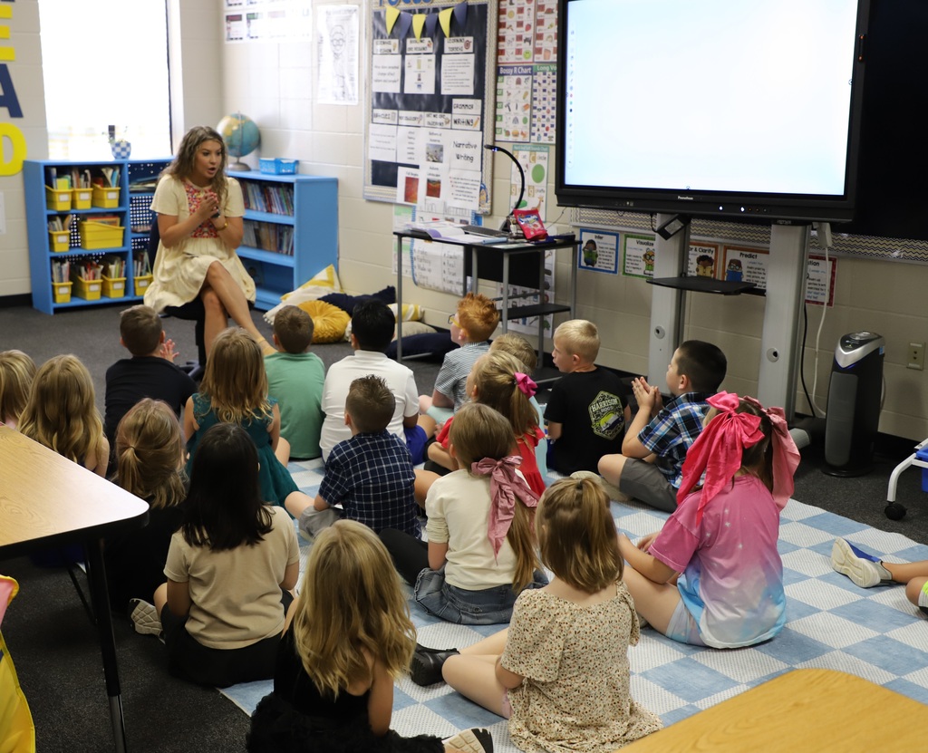 A group of elementary school students sitting cross-legged on a checkered rug, listening attentively to their teacher who is speaking at the front of the classroom.