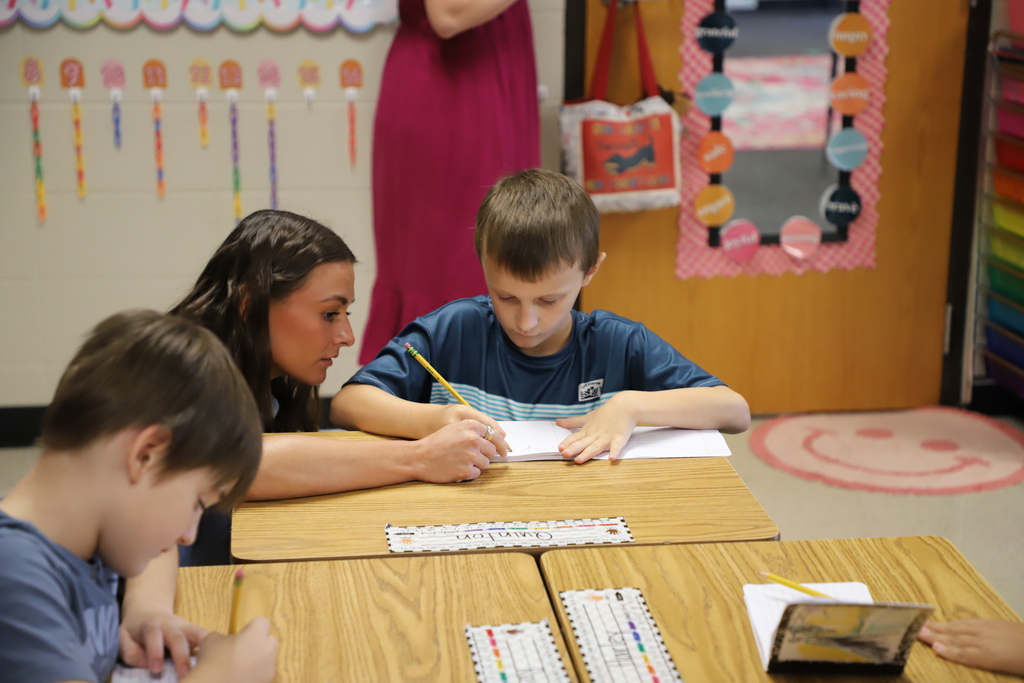 A female teacher leaning in next to a young boy at a school desk, gently guiding his hand as he writes with a pencil in his notebook.