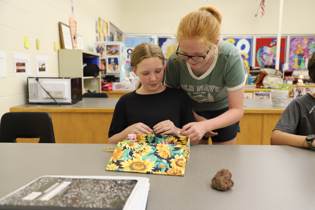 wo young girls in a classroom working together on a hands-on project, looking down attentively at a colorful fabric box patterned with sunflowers on a grey table.