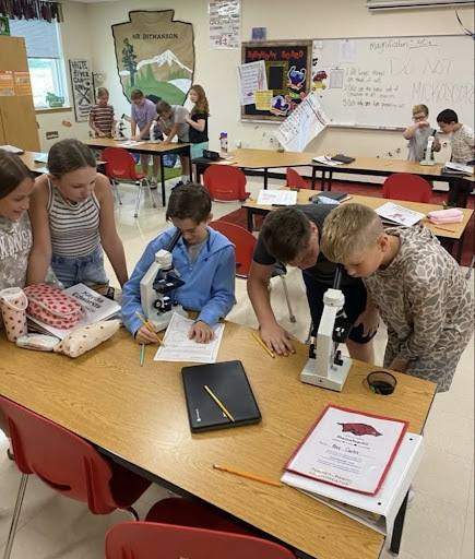 A group of middle school students gathered around a classroom table making homemade ice cream in plastic bags. Two girls in the center measure ingredients, surrounded by salt, sugar, and measuring spoons, while other students assist.