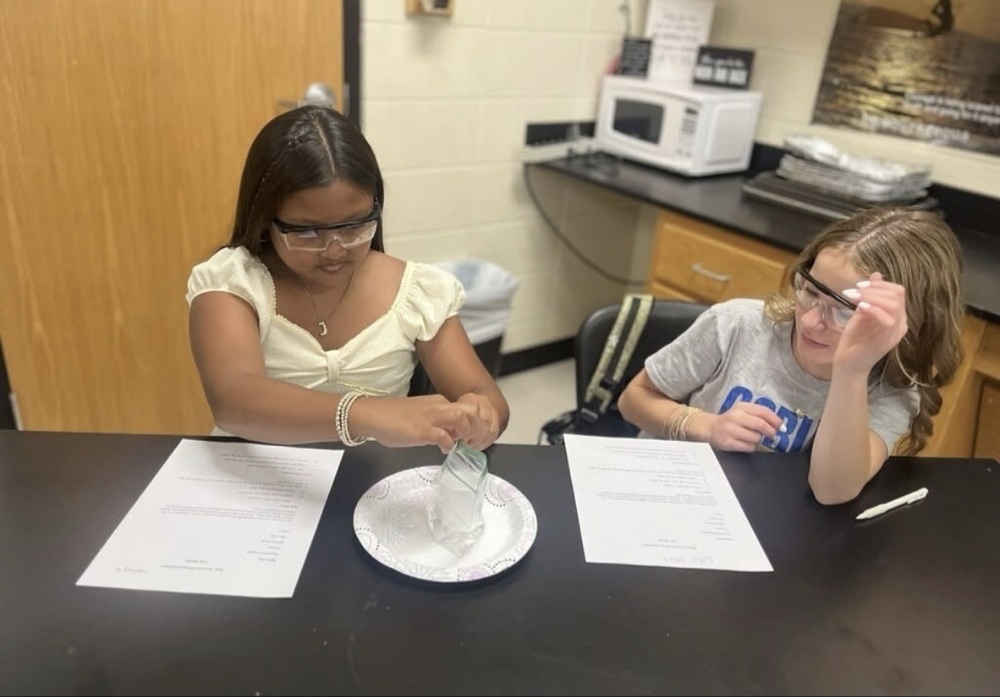 Two middle school girls at a lab bench conduct a science experiment. The student on the left, wearing safety glasses, carefully seals a plastic bag over a paper plate, while her classmate in a gray shirt rests her chin on her hand and observes the steps.