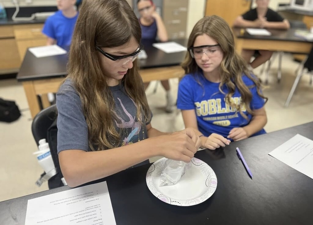 Two Harrison Middle School girls wearing safety glasses sit at a lab table. One girl in a gray t-shirt pours baking soda into a plastic bag over a paper plate, following instructions on a worksheet, while her teammate in a blue "Goblins" t-shirt watches closely.