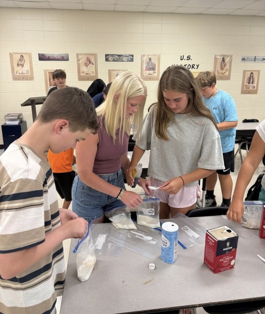 A group of middle school students gathered around a classroom table making homemade ice cream in plastic bags. Two girls in the center measure ingredients, surrounded by salt, sugar, and measuring spoons, while other students assist.