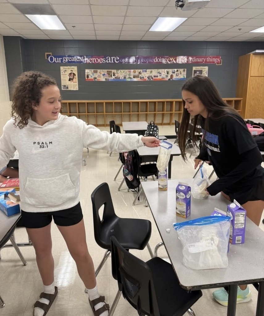Two middle school girls in a classroom work on an experiment together at a table. One student in a white hoodie holds up a clear plastic gallon bag containing a white mixture, while her classmate in a black long-sleeve shirt adds milk or cream from a carton.