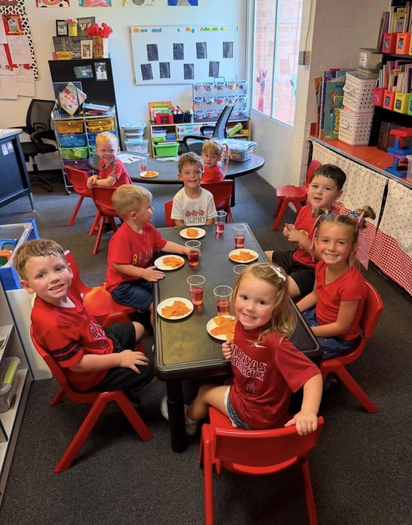 Seven preschool children wearing red shirts sit around a kidney-shaped black table and a small red table in a colorful classroom. They are smiling at the camera, with plates of orange cracker snacks and cups of red juice in front of them on the tables.
