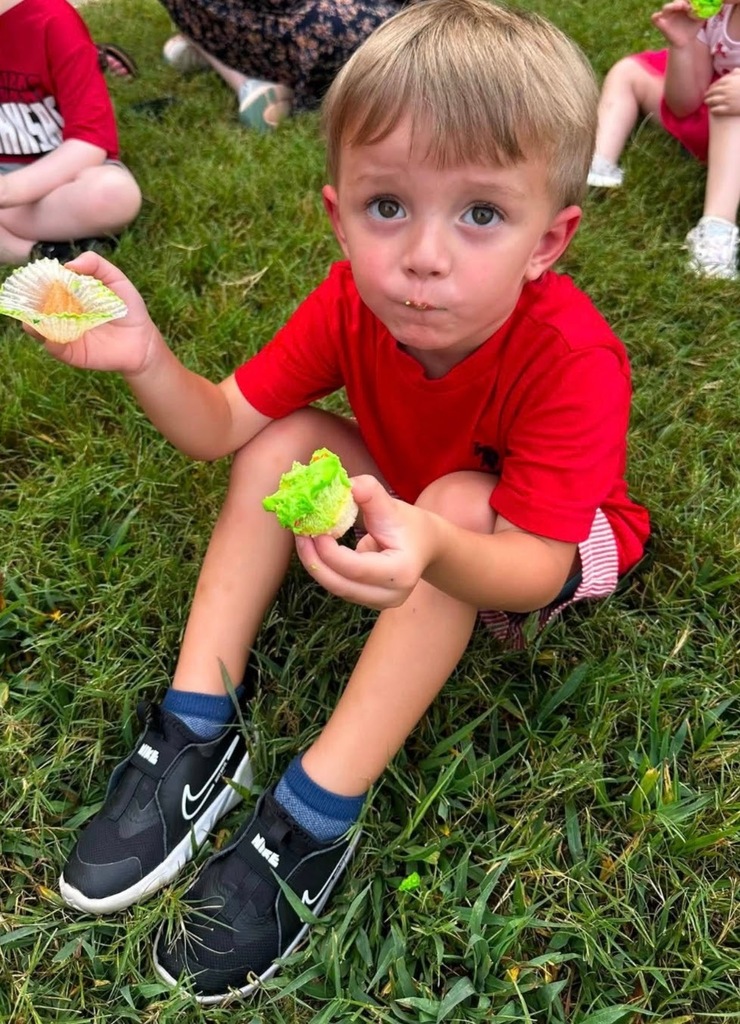 A young preschool boy wearing a bright red t-shirt sits on the green grass outside. He is holding a half-eaten cupcake with bright green frosting in one hand and the empty cupcake wrapper in the other while looking directly at the camera. Other children in red shirts can be seen sitting on the grass nearby in the background.
