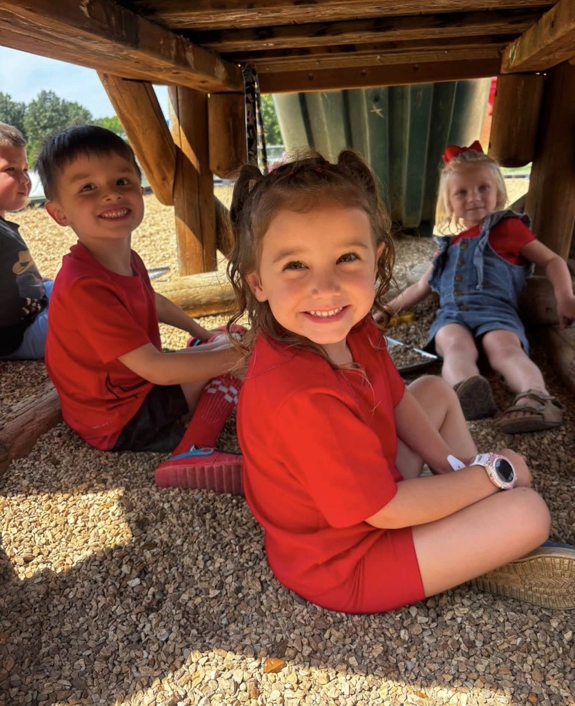 Three preschool children, two girls and a boy, sit together smiling underneath a wooden playground structure on a pebble floor. They are all wearing red shirts, and the girl in the background wears a large red bow.