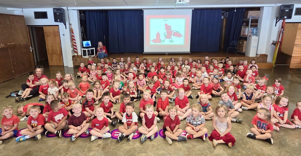 A large group of preschool children, nearly all wearing red shirts, sit cross-legged on the floor of a school gymnasium or auditorium for an assembly. On the stage behind them, a projector screen displays a cartoon red crayon and the word "Red." Two adult teachers sit on the sides supervising the large group.